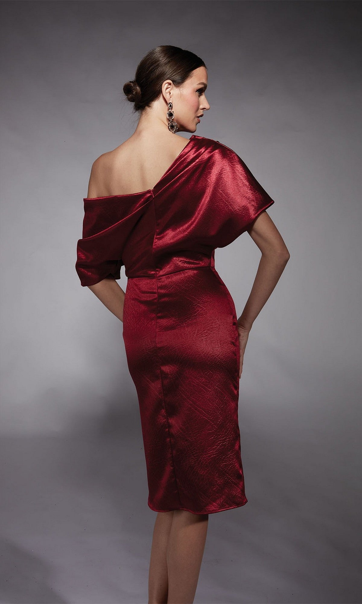 A woman stands in a studio facing away, wearing the Alyce 70060 knee-length semi-formal dress in shiny burgundy charmeuse with an off-shoulder design; her hair is styled in a low bun.